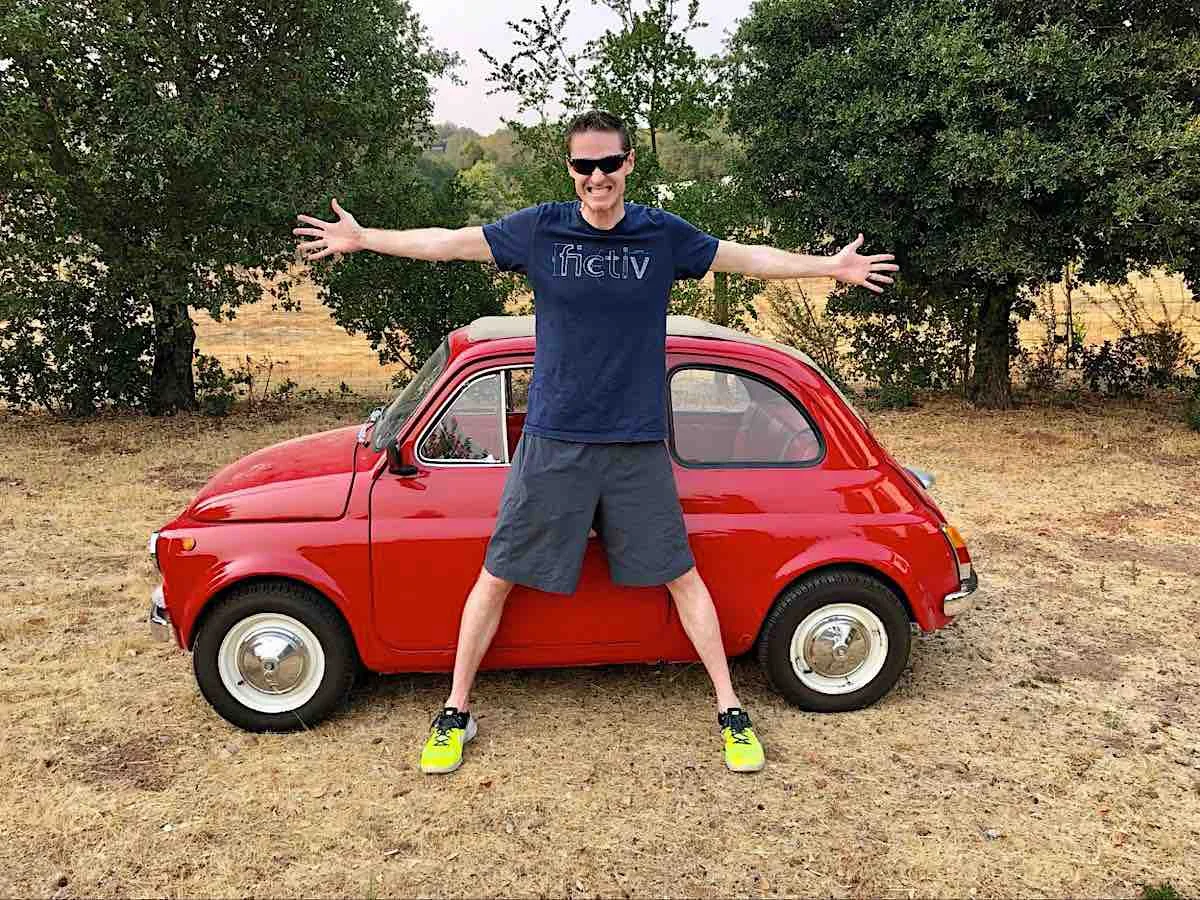 Image of a bright red fiat 500 with a tall man standing in front of it with his arms stretched out to either side and legs wide apart