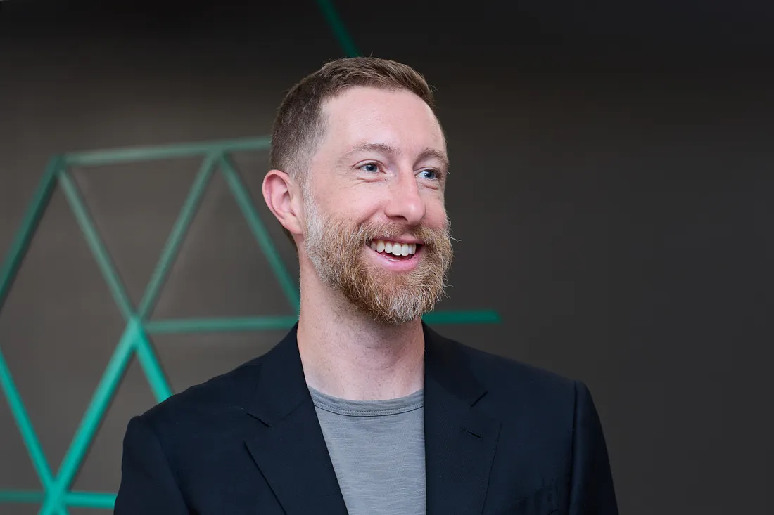 Smiling man with short hair and beard in a dark blazer, gray tee, standing in front of teal geometric art.