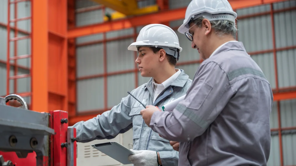 Two factory workers wearing hard hats inspect a machine, one holds a clipboard while the other points and discusses details