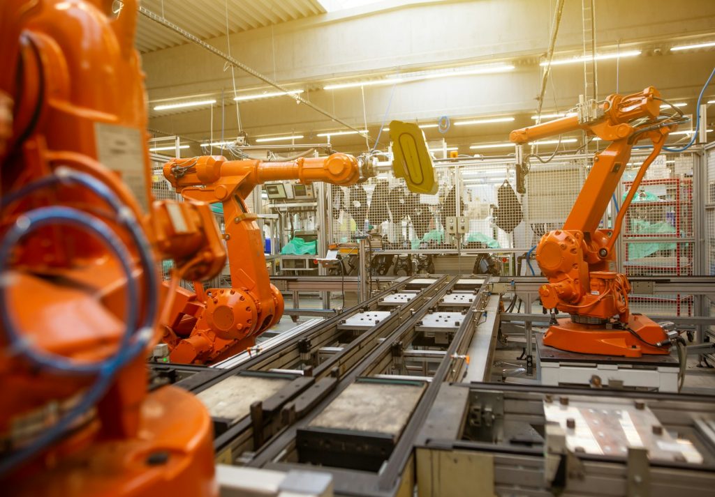 Orange robotic arms operate over a metal conveyor on a bright factory assembly line.