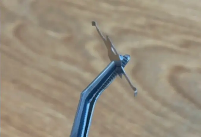thin-metal-parts-img-1 - Fictiv Tweezer holding a small metal cross-shaped component above a wooden surface.