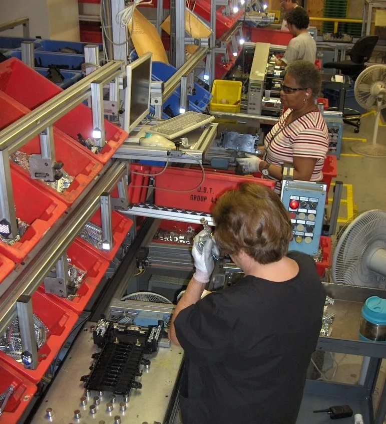 Workers on an electronics assembly line placing components into red bins and operating a benchtop station with a control panel and computer screen nearby