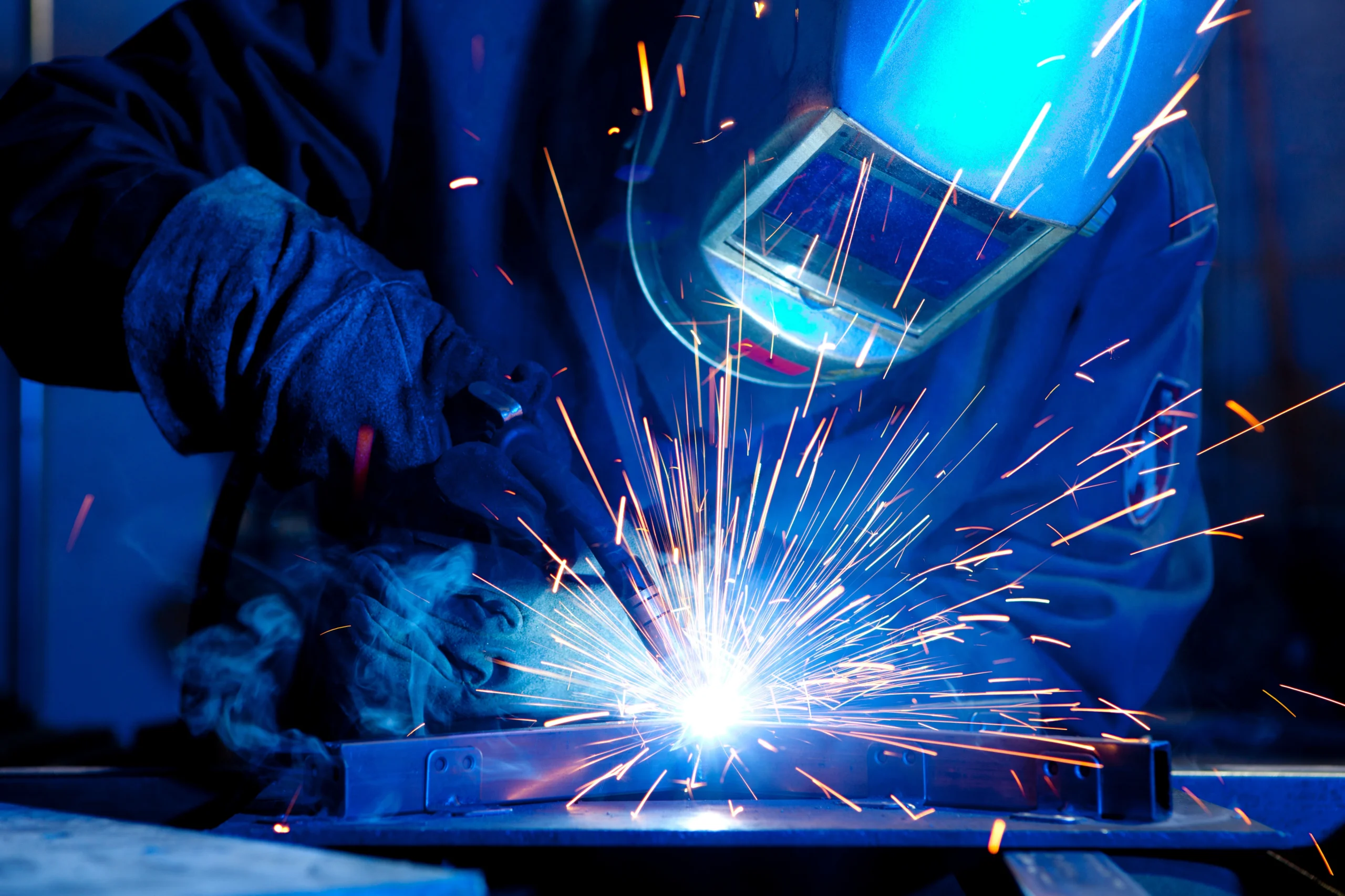 Welding a metal piece with bright sparks and blue protective gear visible in a dark workshop.