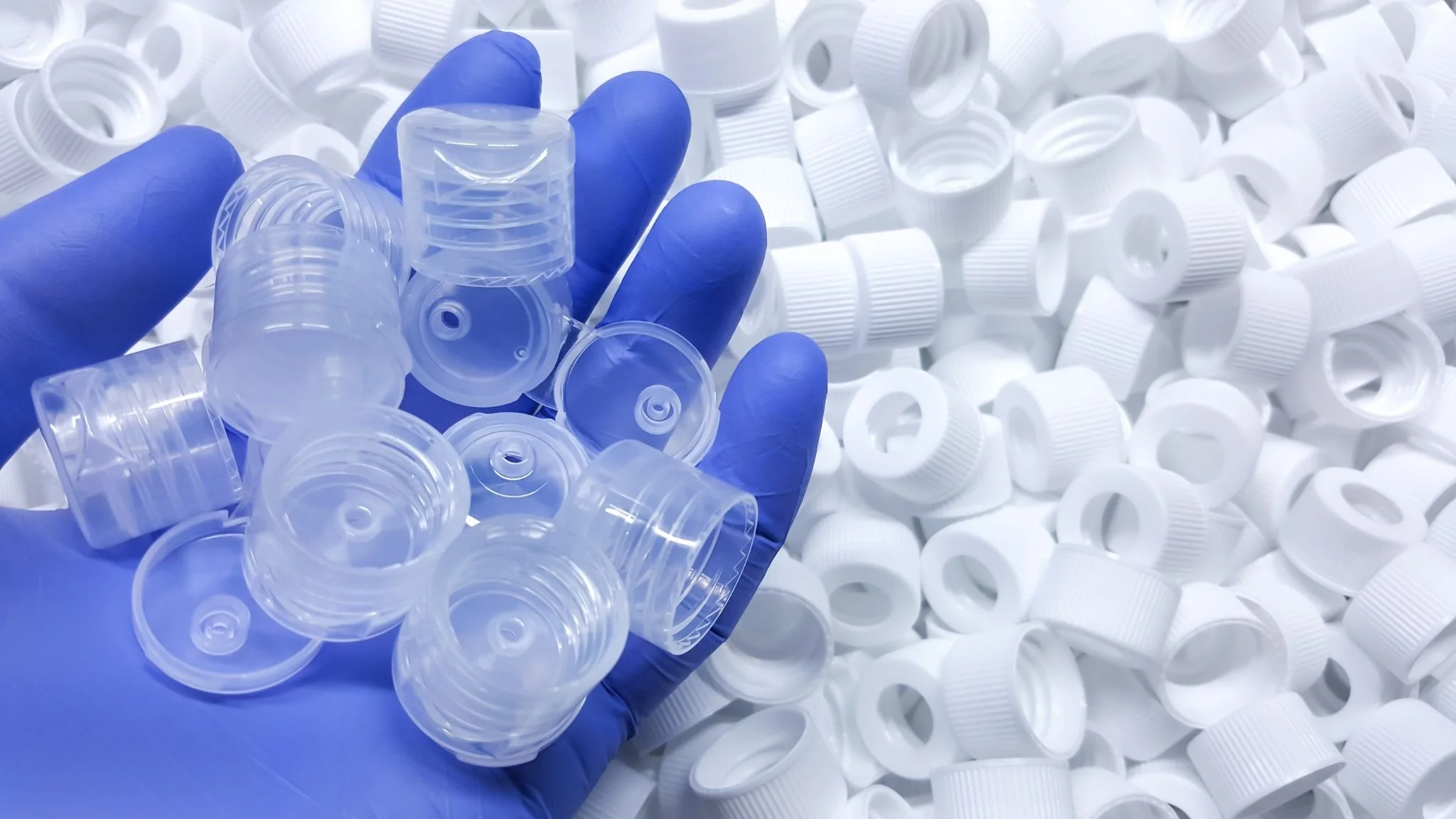 Gloved hand holding several clear plastic bottle caps over a pile of white caps, close-up scene for recycling or lab work.