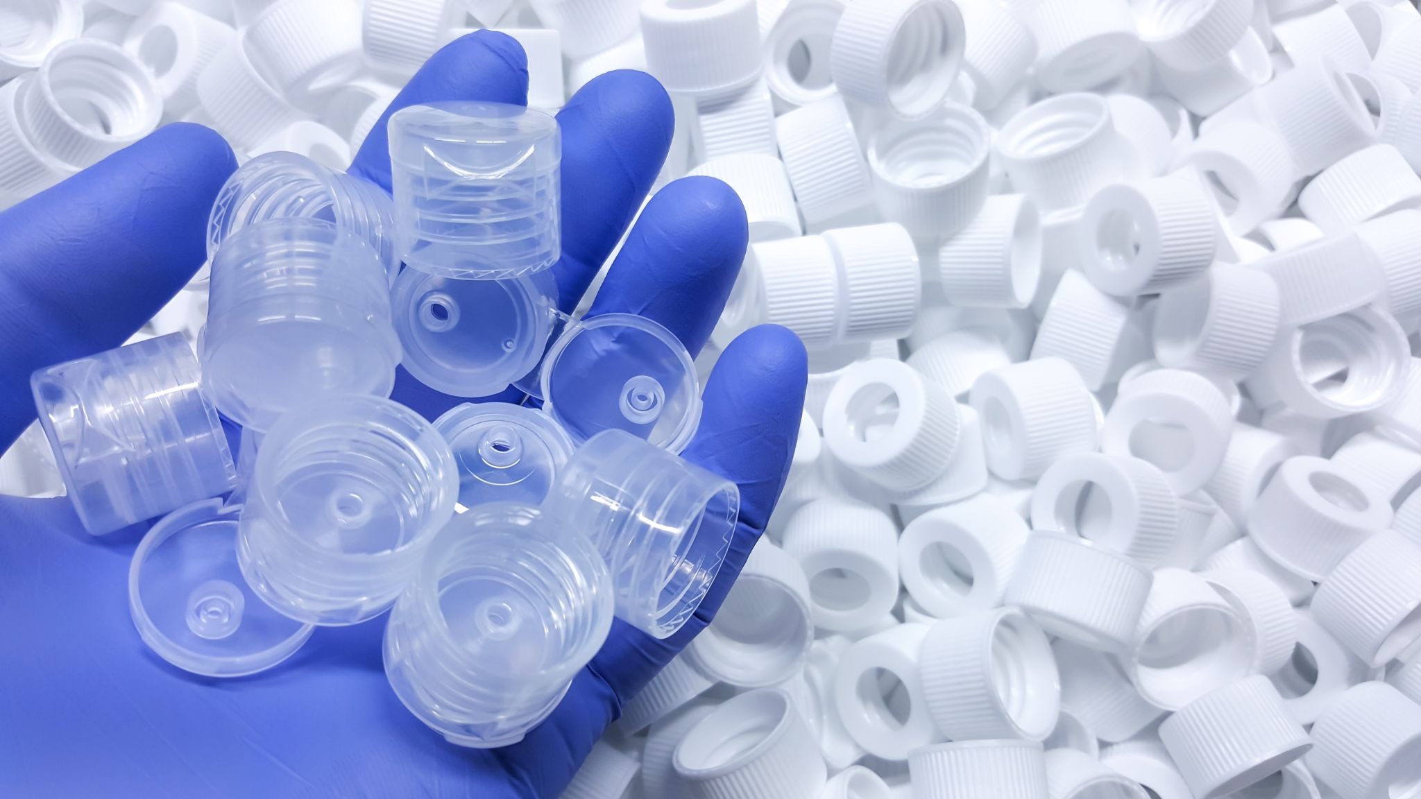 image - Fictiv Gloved hand holding several clear plastic bottle caps over a pile of white caps, close-up scene for recycling or lab work.