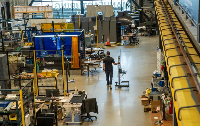Man walking down a spacious factory floor past workstations and industrial equipment, with yellow safety rails along a mezzanine.