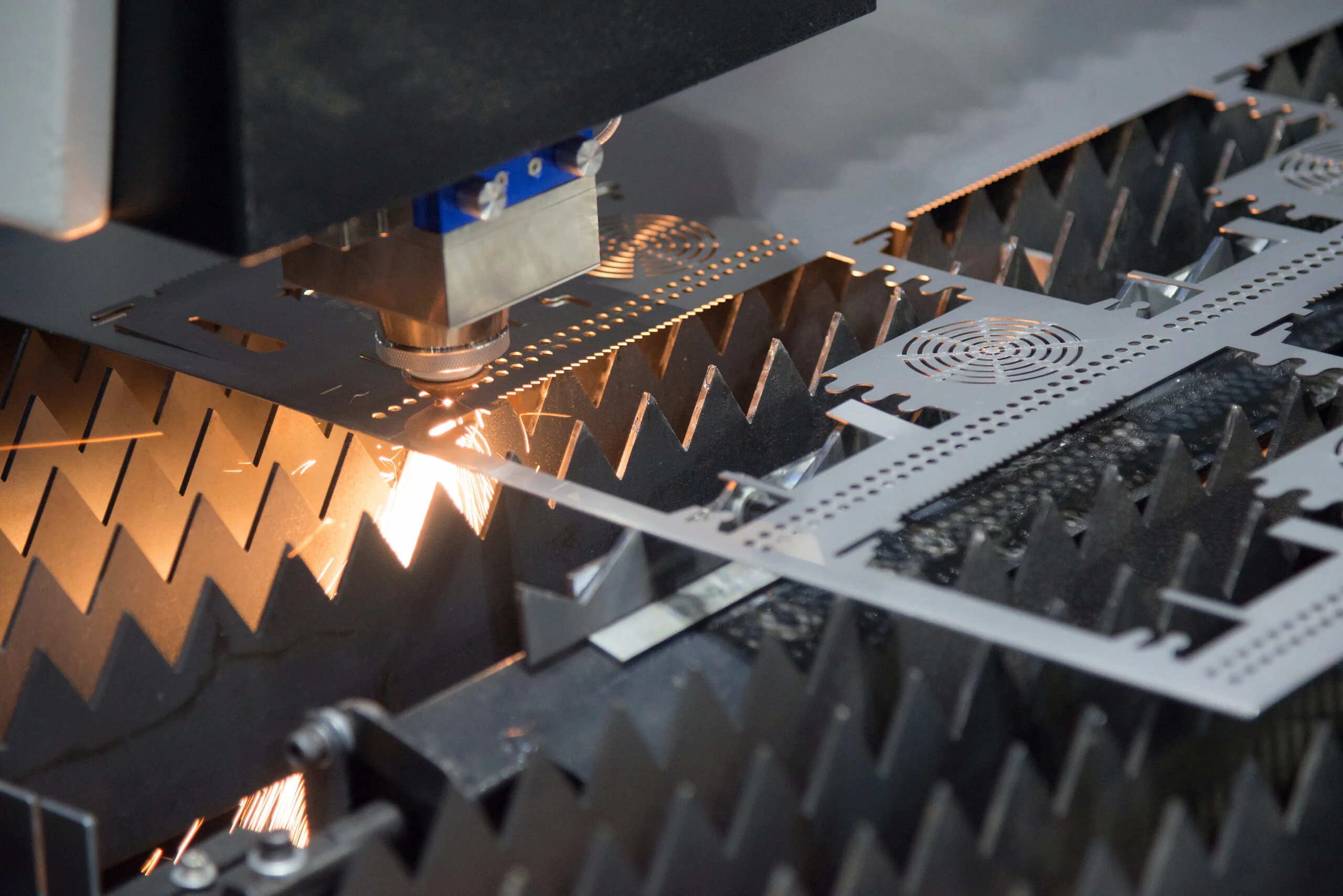 The laser cutter machine while cutting the sheet metal with the sparking light - Fictiv Laser cutting machine cutting a metal sheet, sparks flying in a dark industrial setting.